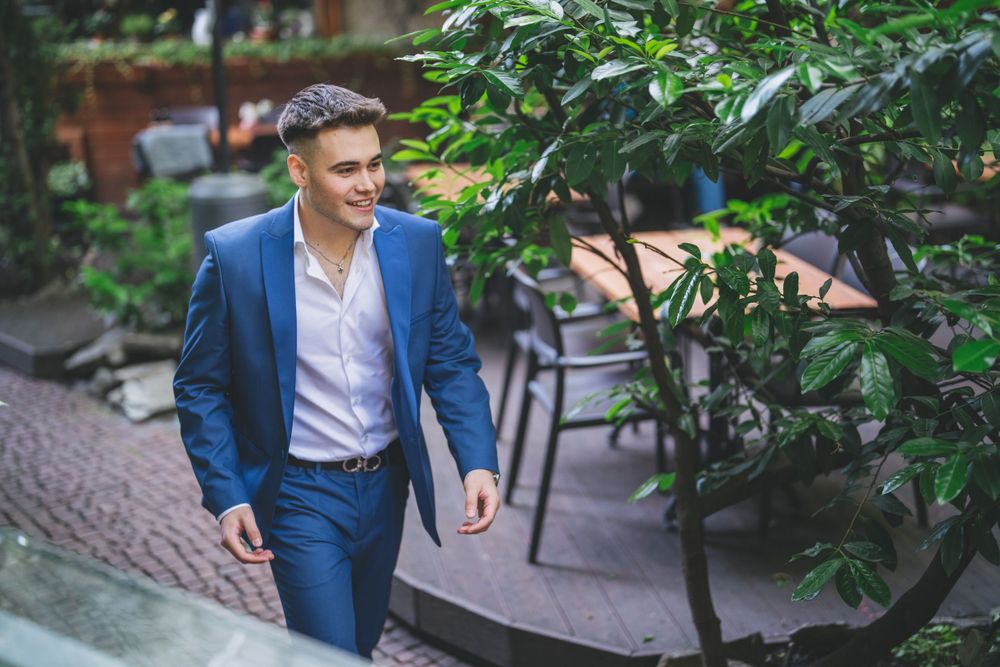 Person in blue suit walks along garden path with greenery and brick pavement.