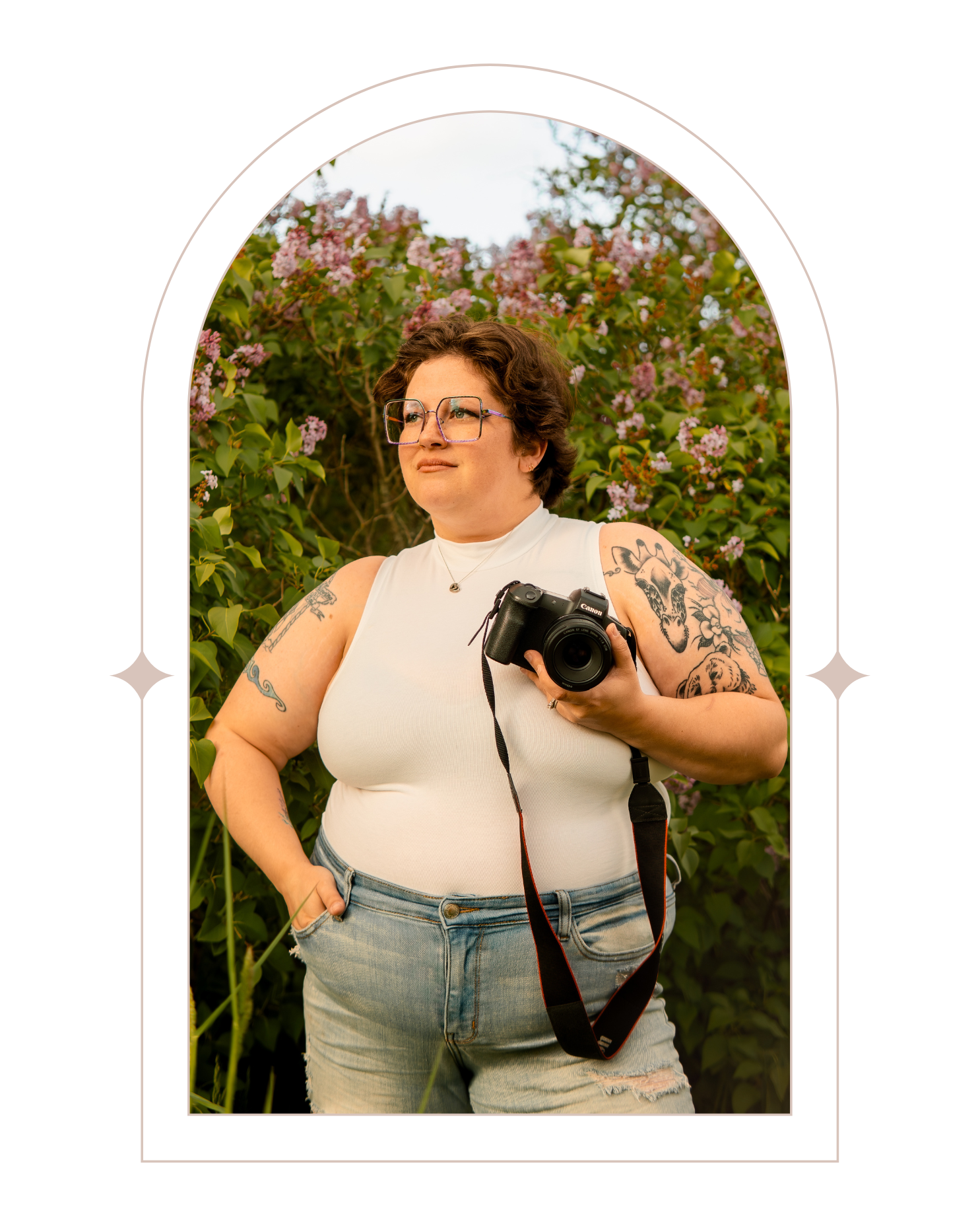 A photographer holding a DSLR camera stands in front of blooming flowers wearing a white tank top and jeans.