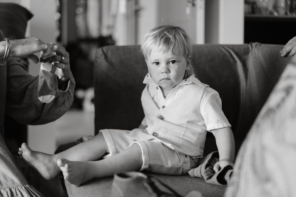 A young child in white sits on a dark leather couch during a casual gathering.