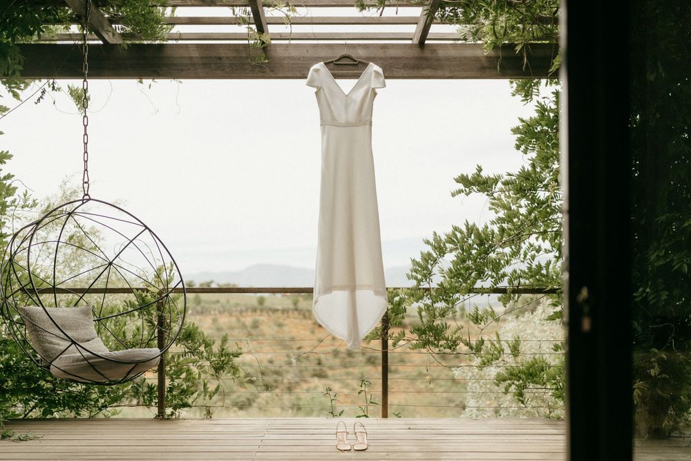 White wedding dress hanging elegantly against a bright background with greenery framing the scene.