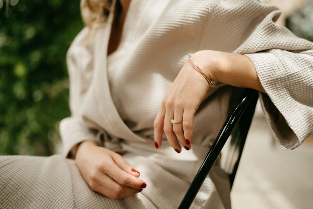 Close-up view of hands folded in lap wearing beige knit sweater while seated on a chair with greenery in background.