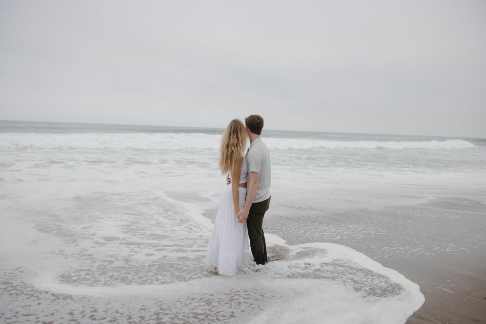 A couple stands together at the ocean's edge as waves gently lap at their feet on a misty beach.