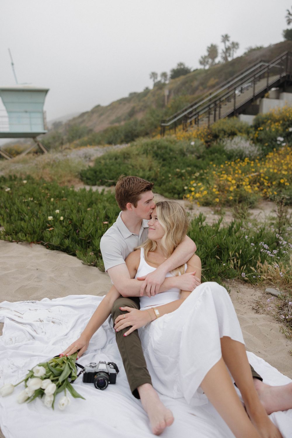 A couple sits together on a white blanket near a beach path with flowers and a vintage camera nearby.