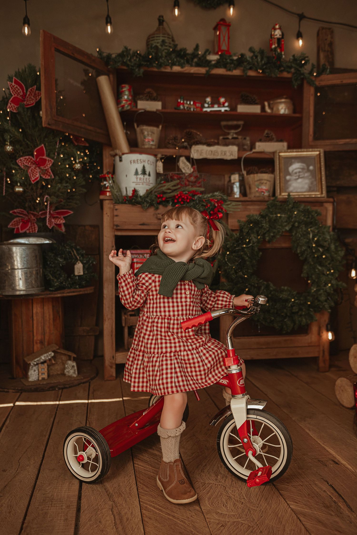 A child on a red tricycle plays in a rustic holiday-decorated room with twinkle lights and garland.