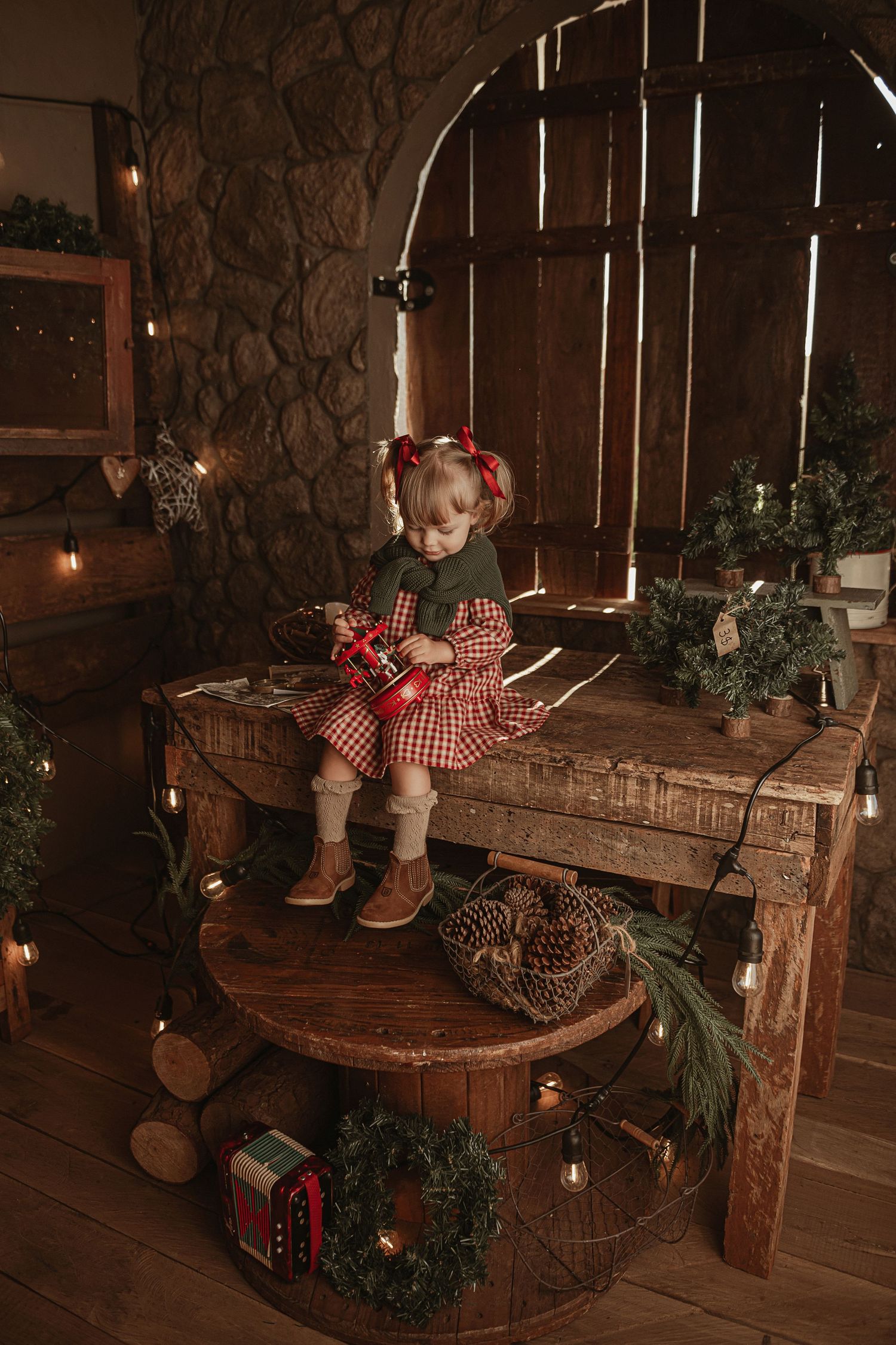 Cozy rustic Christmas scene with wooden furniture, plaid decor, wreaths and twinkling lights in a barn-like setting.