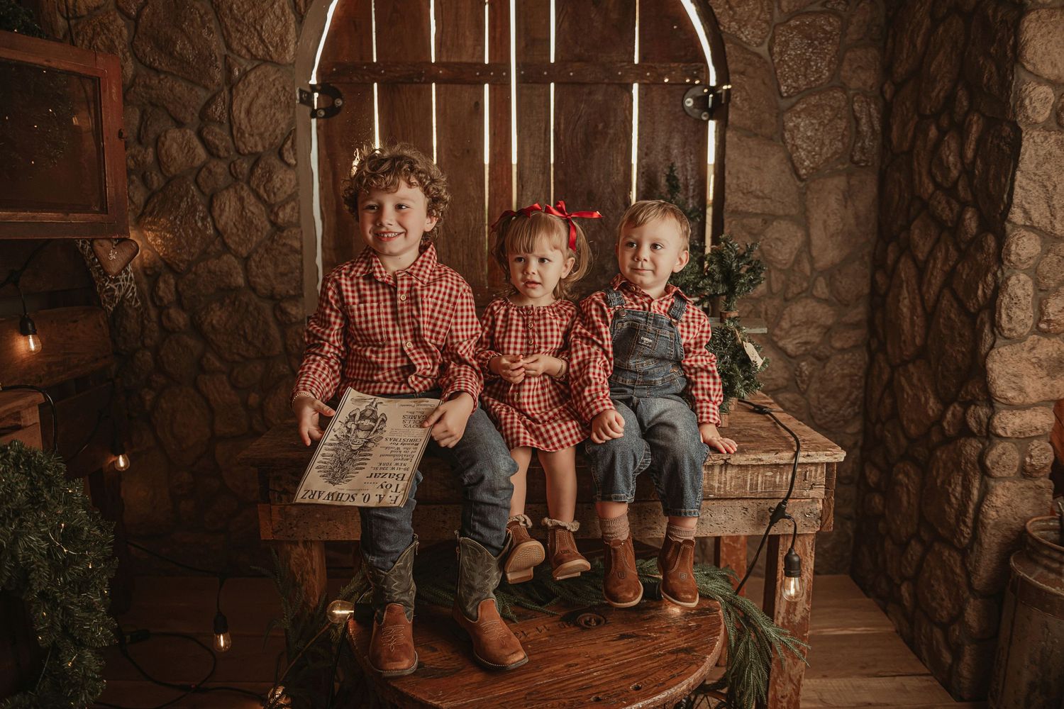 Three children in matching plaid shirts sit together on a rustic wooden bench in a stone-walled room with twinkling lights.