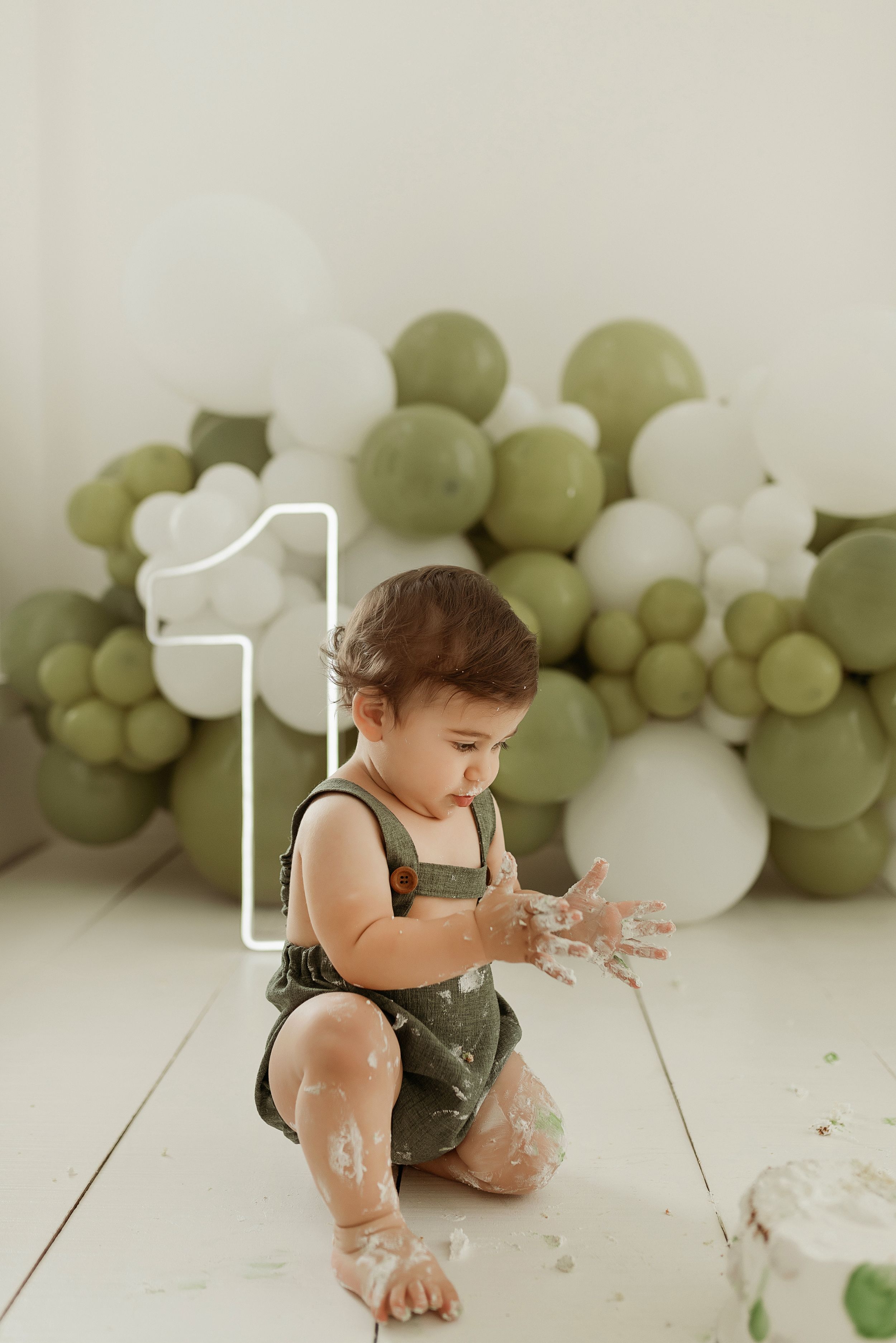Baby in olive overalls sitting on floor with green and white balloon arch backdrop and number 1 decoration.