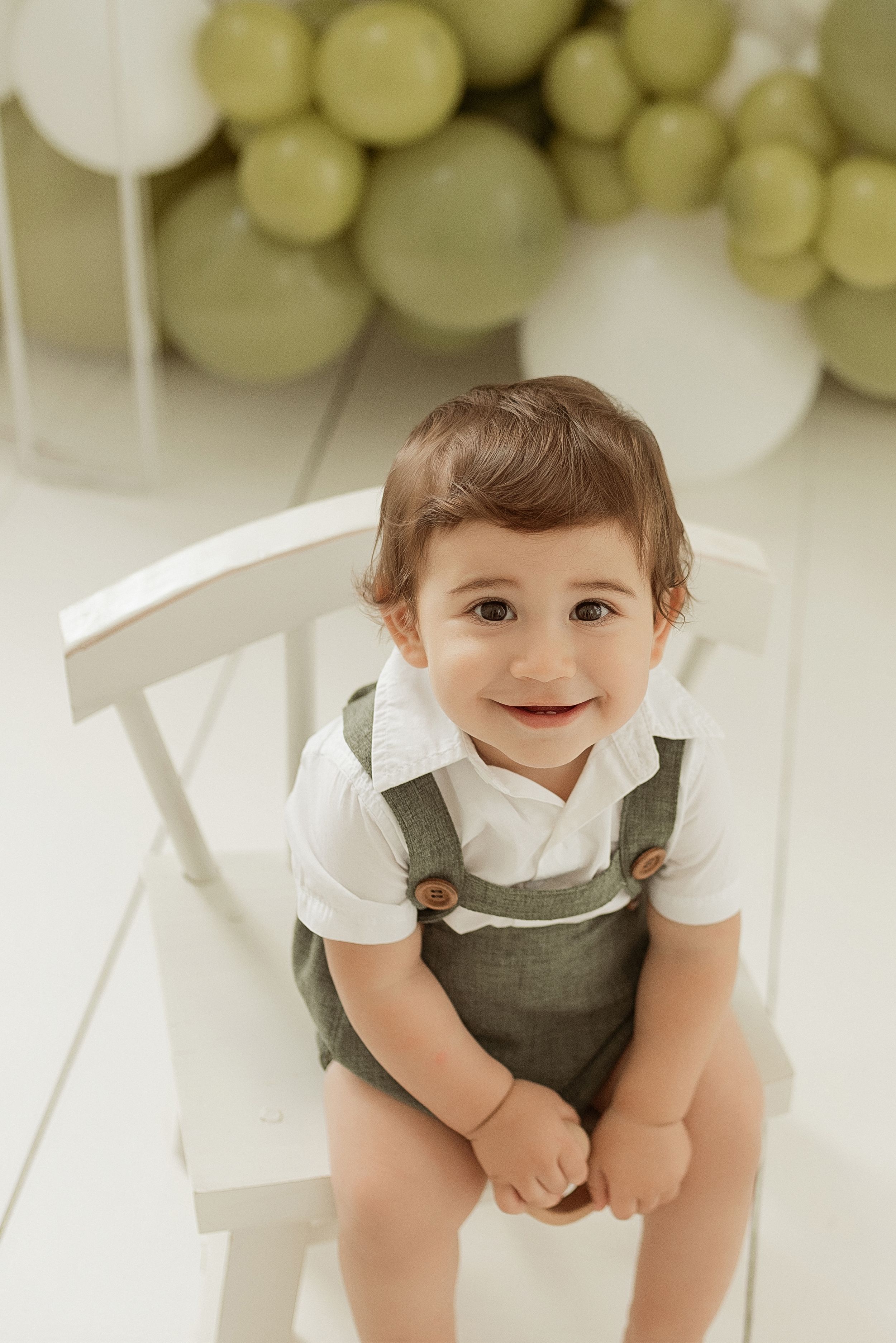 A small child wears olive green overalls and a white shirt while sitting on a white chair with balloon decor in background.