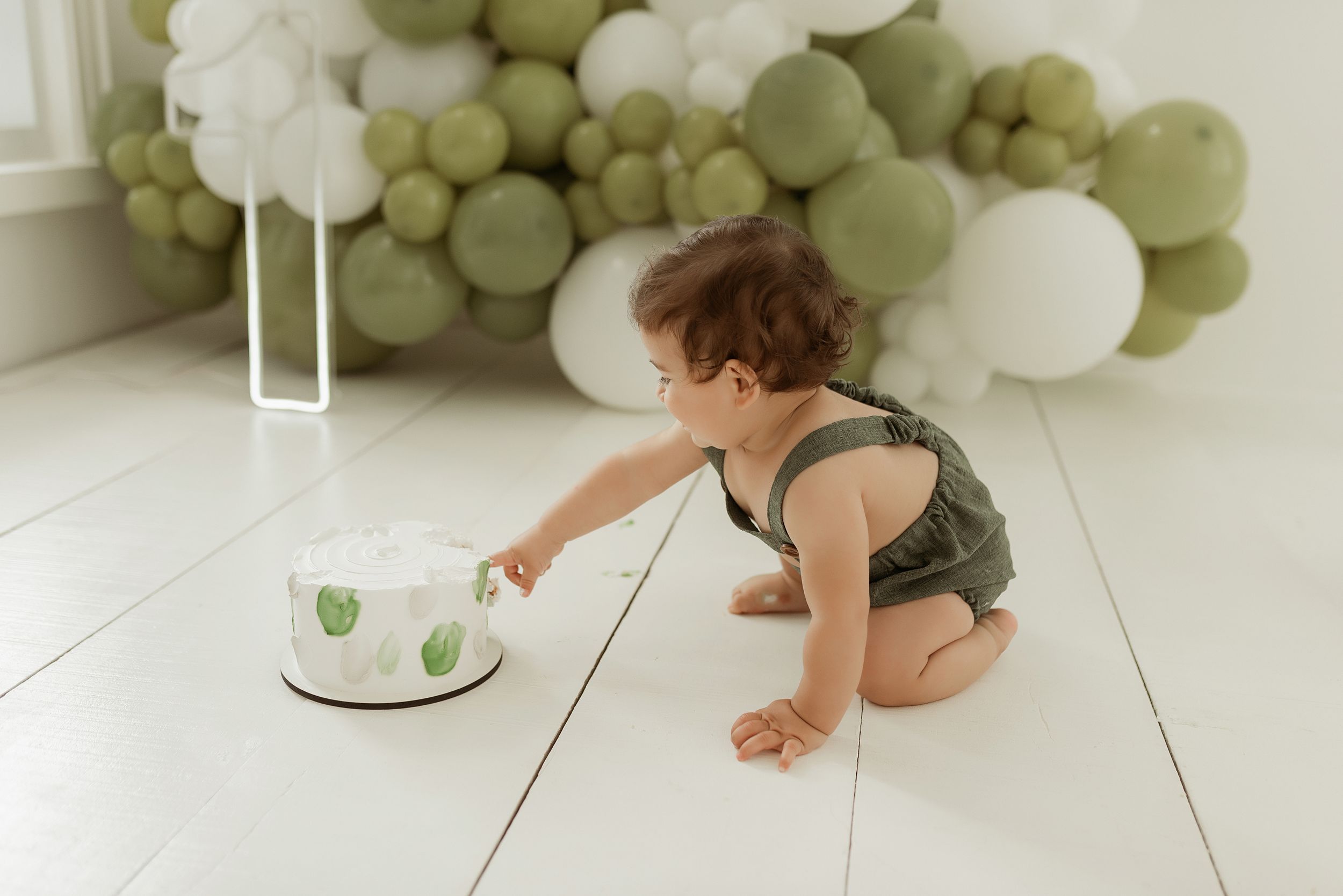 A small child in olive romper crouches to inspect a cake with green decorations against a backdrop of sage and white balloons.