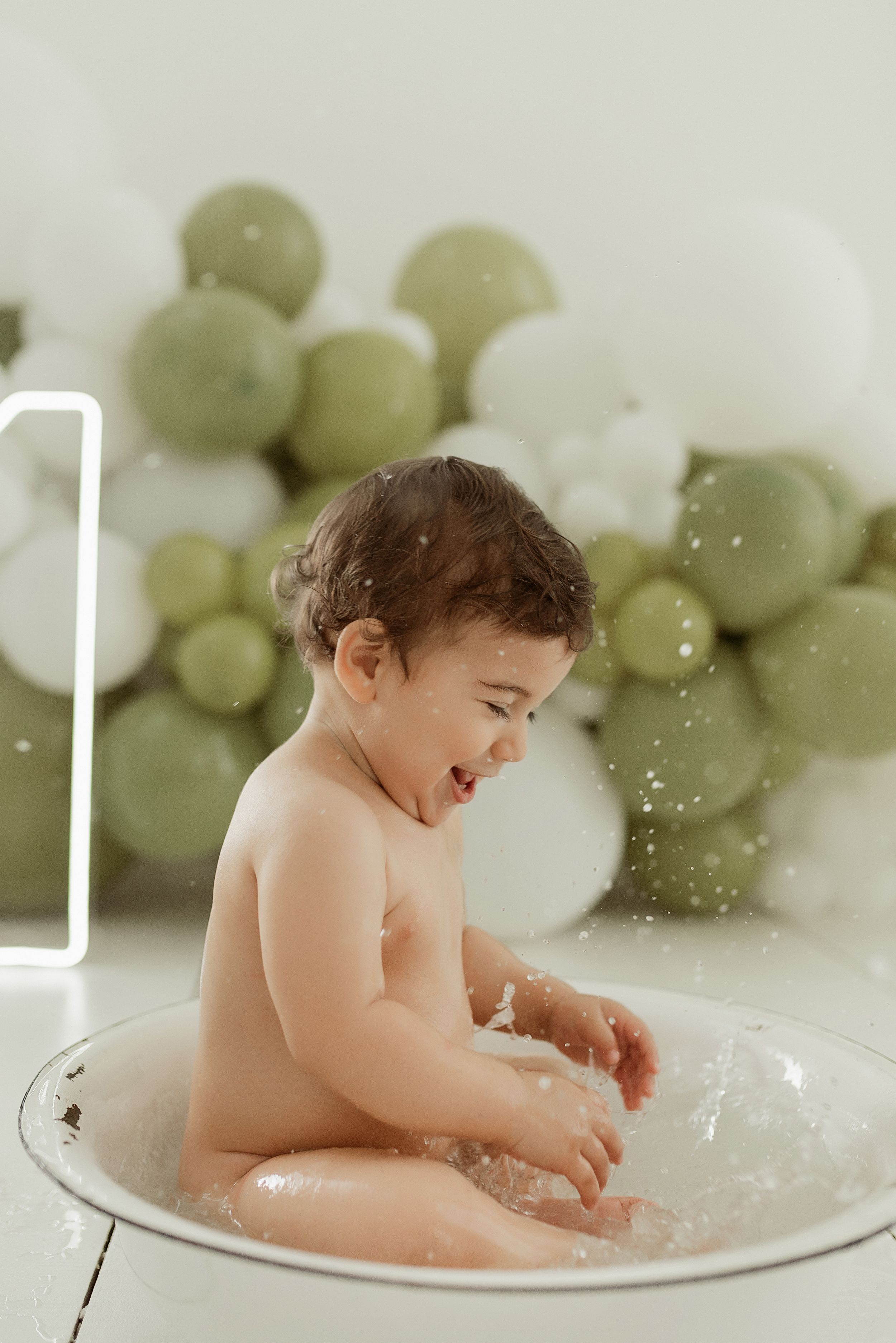 Child laughs joyfully during bath time with green balloon decorations in background.