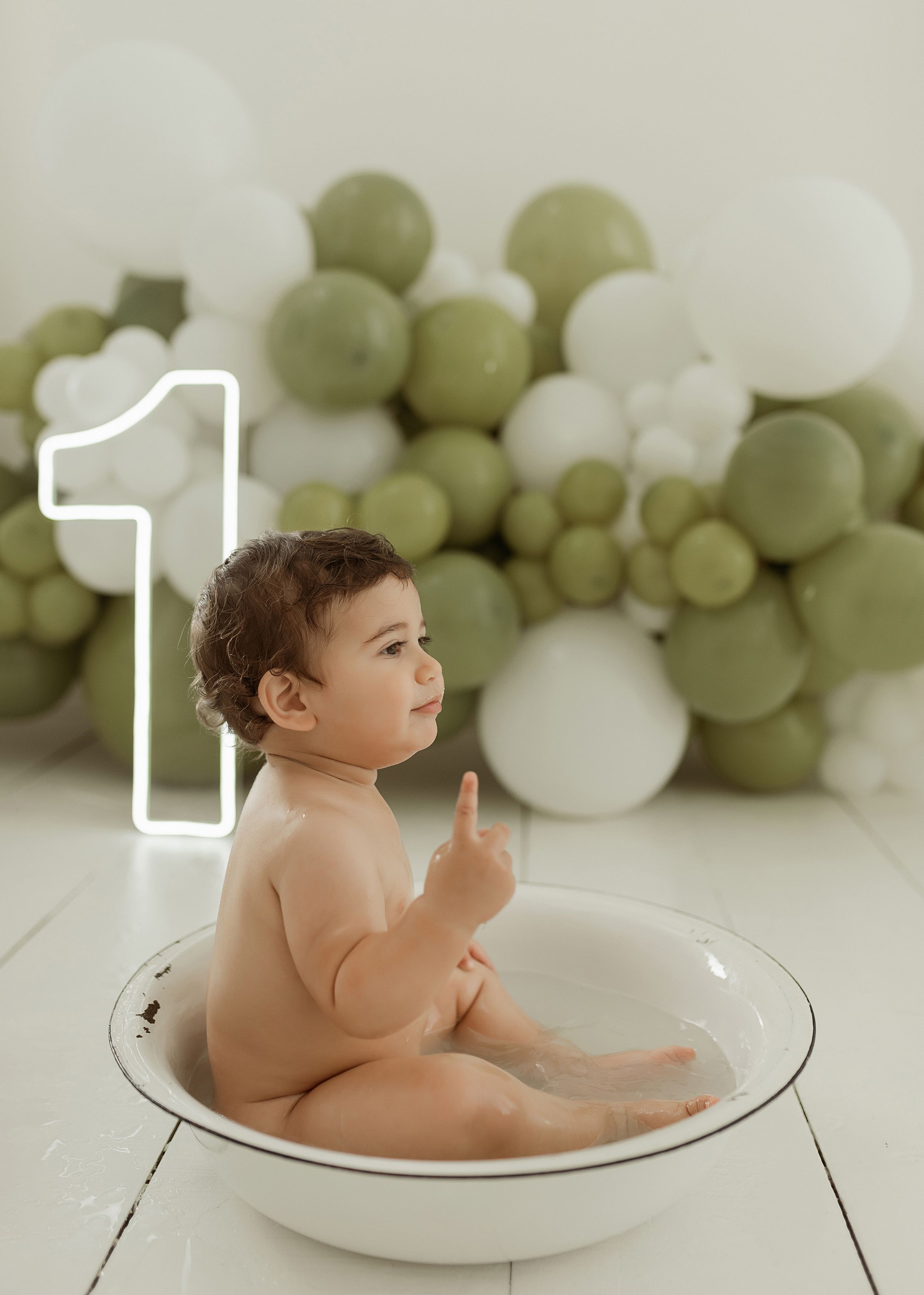 A baby sits in a white basin with a balloon arch display featuring green and white balloons and a large number one.