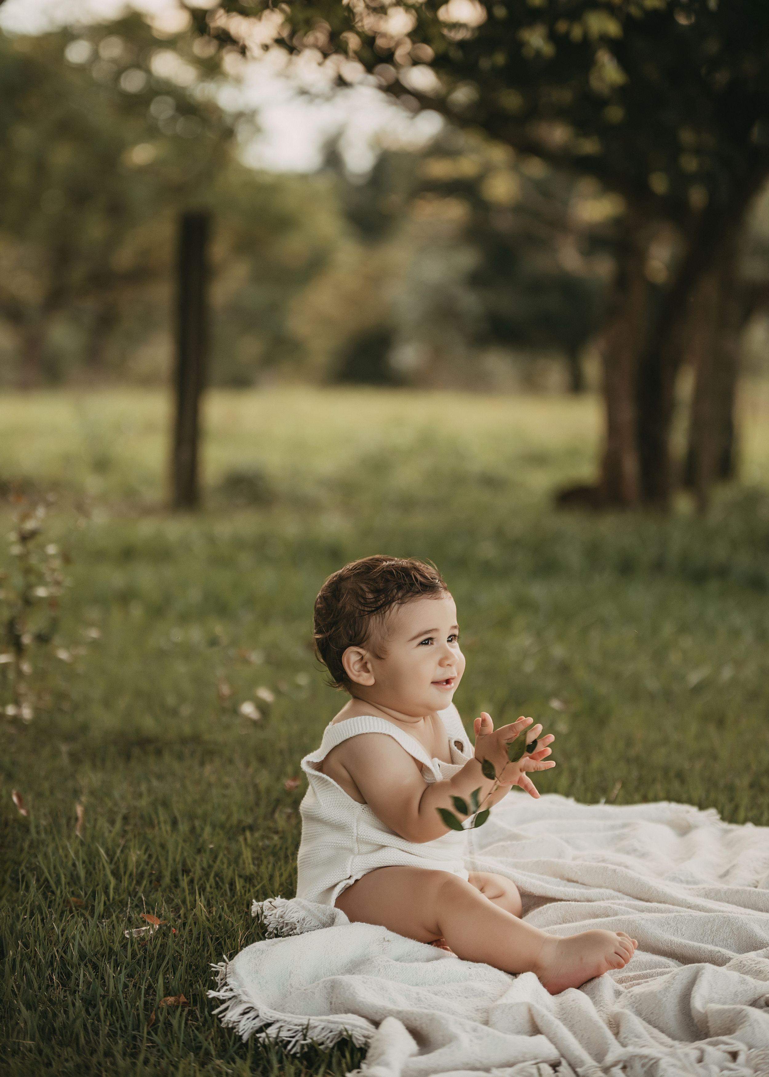 Small child in white dress sits on cream blanket in dreamy outdoor garden setting during golden hour.