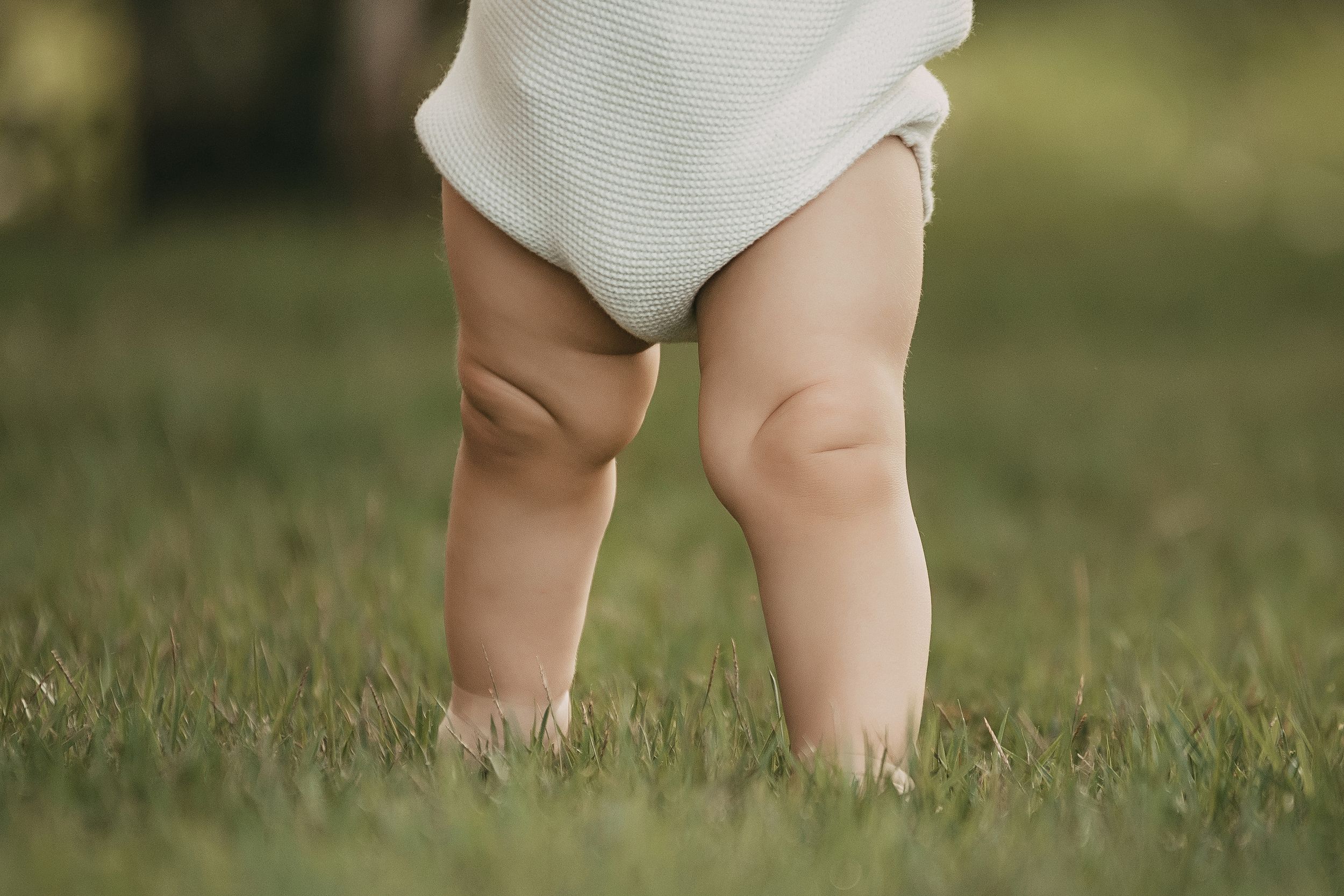 Close-up view of chubby baby legs standing barefoot in green grass wearing a white romper.