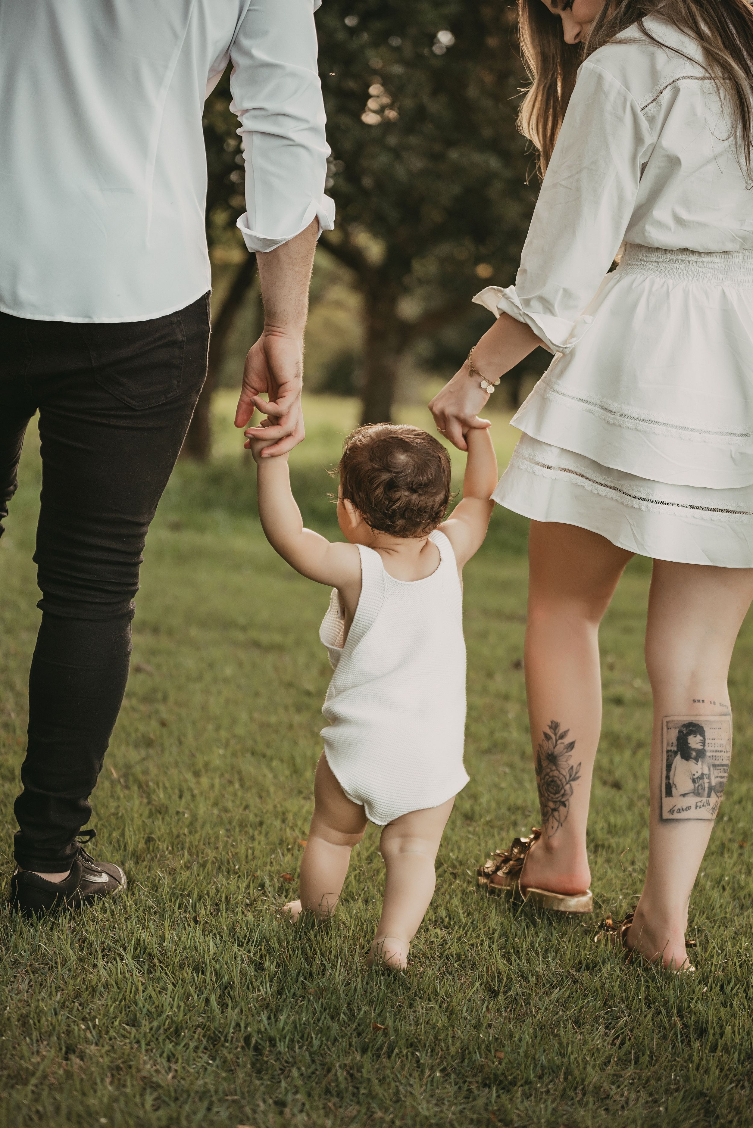 A small child in white holding hands with two others walks on green grass during a sunny summer day.