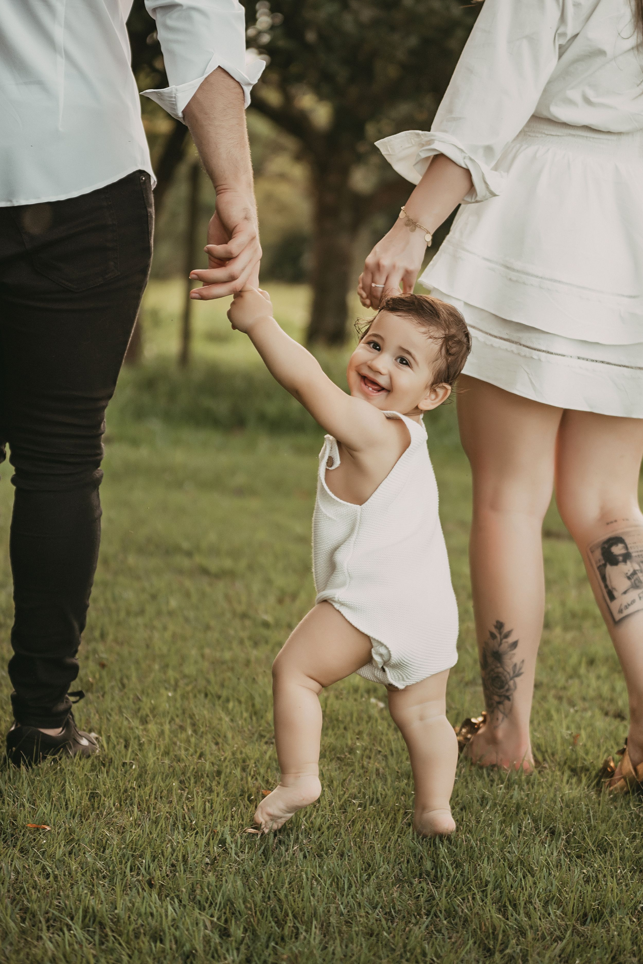 A toddler in white takes first steps on grass while holding parents' hands in a candid outdoor moment.