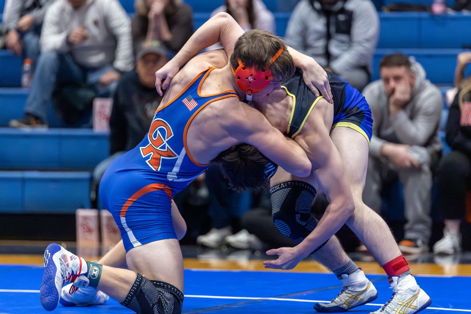 Wrestling match in progress on blue mats with competitors engaged in ground positions.