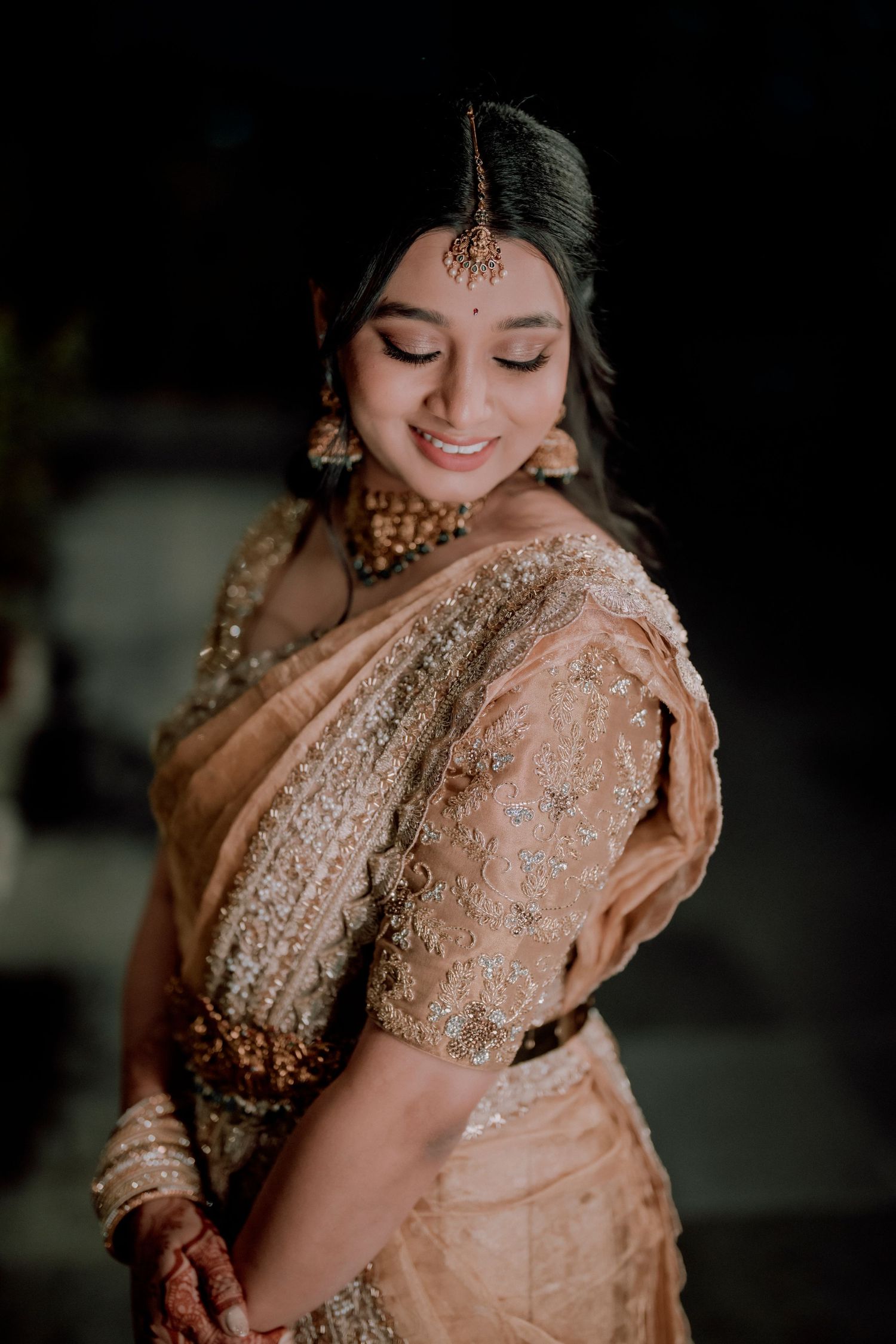 Bride in peach embroidered saree and traditional jewelry smiles with eyes closed.