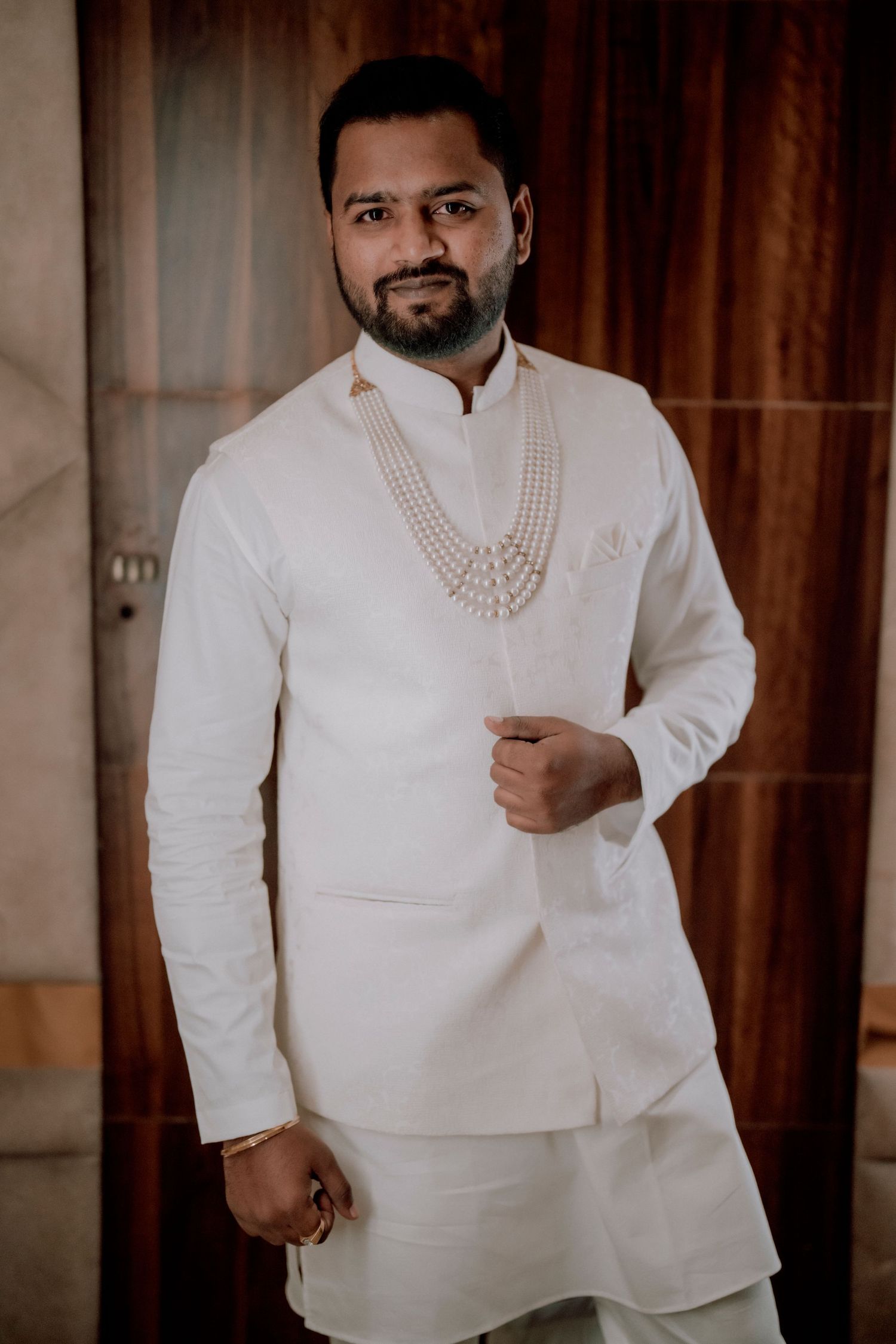 Groom wearing an elegant white Indian wedding outfit with a pearl necklace against a warm backdrop.