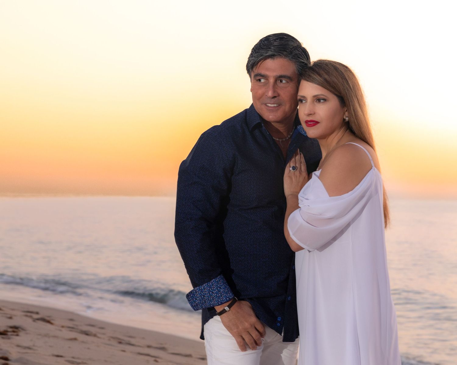 Couple in white and navy blue embrace on beach during golden sunset.