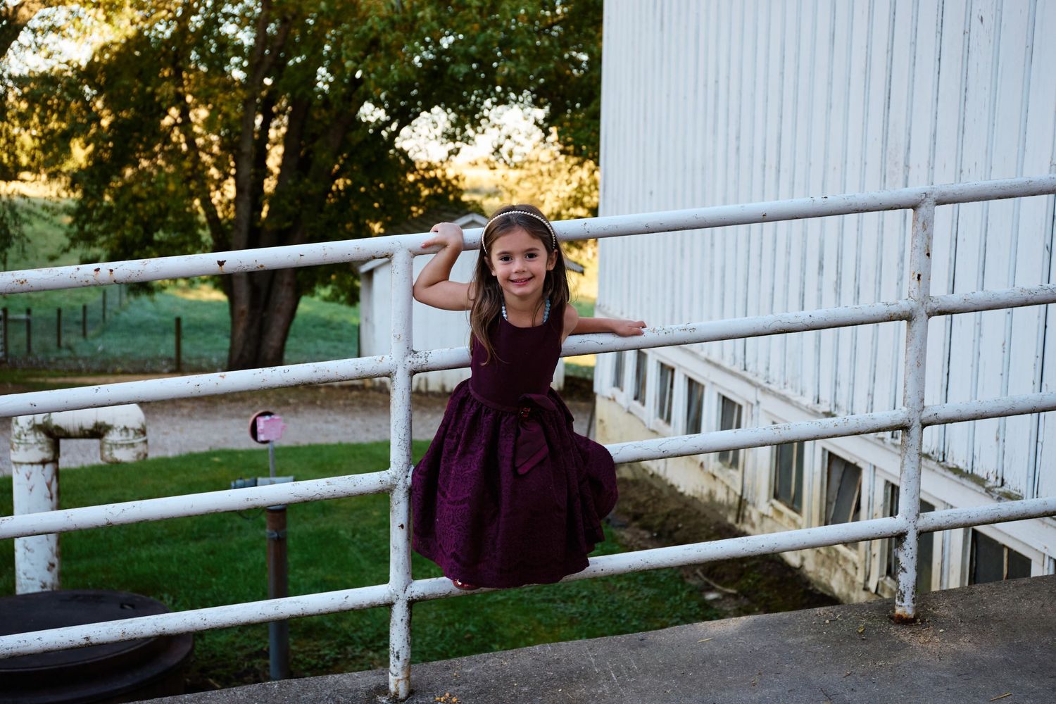 Girl in purple dress playing on white railing near rustic barn.