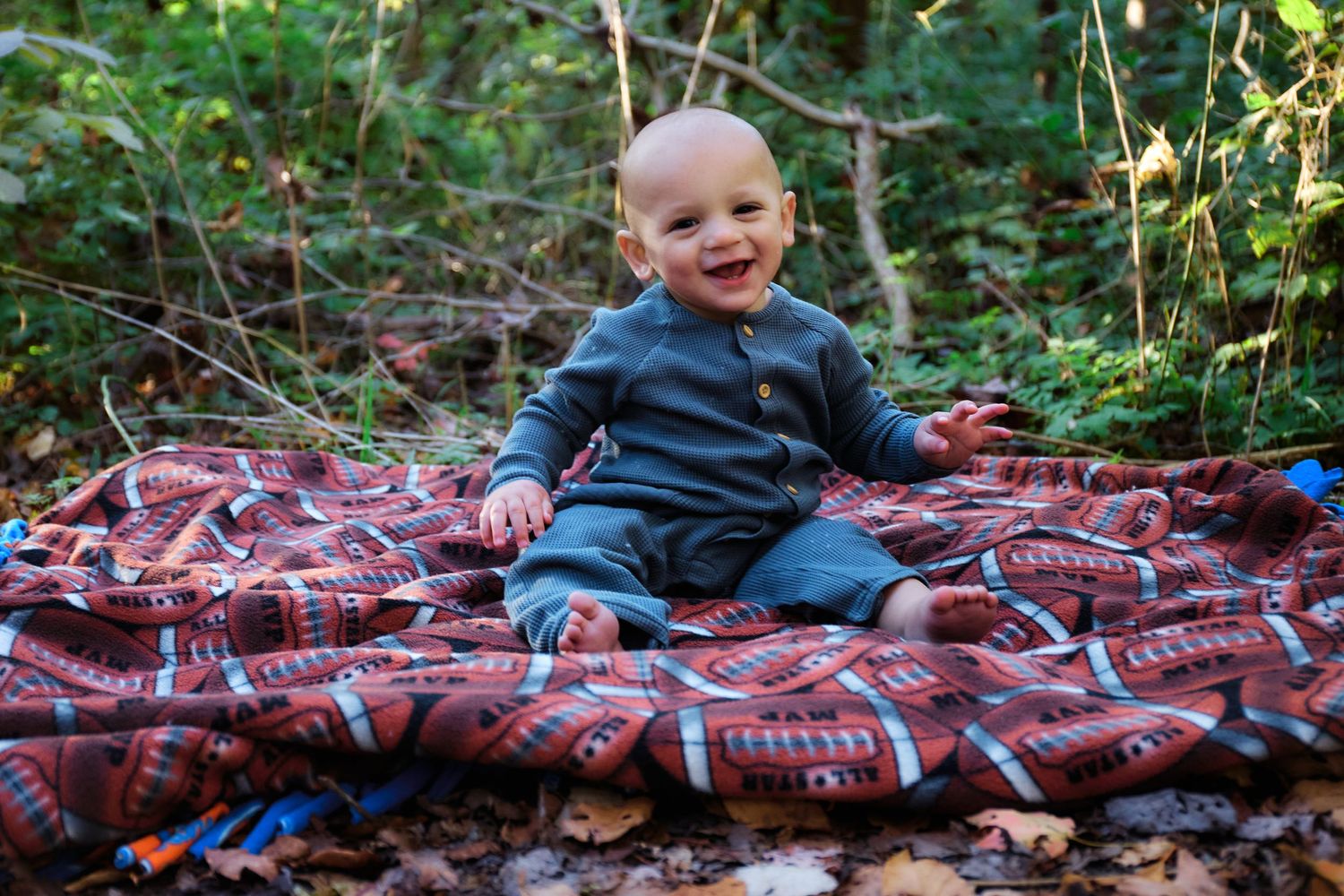 Baby in navy outfit sitting on patterned blanket outdoors while smiling.