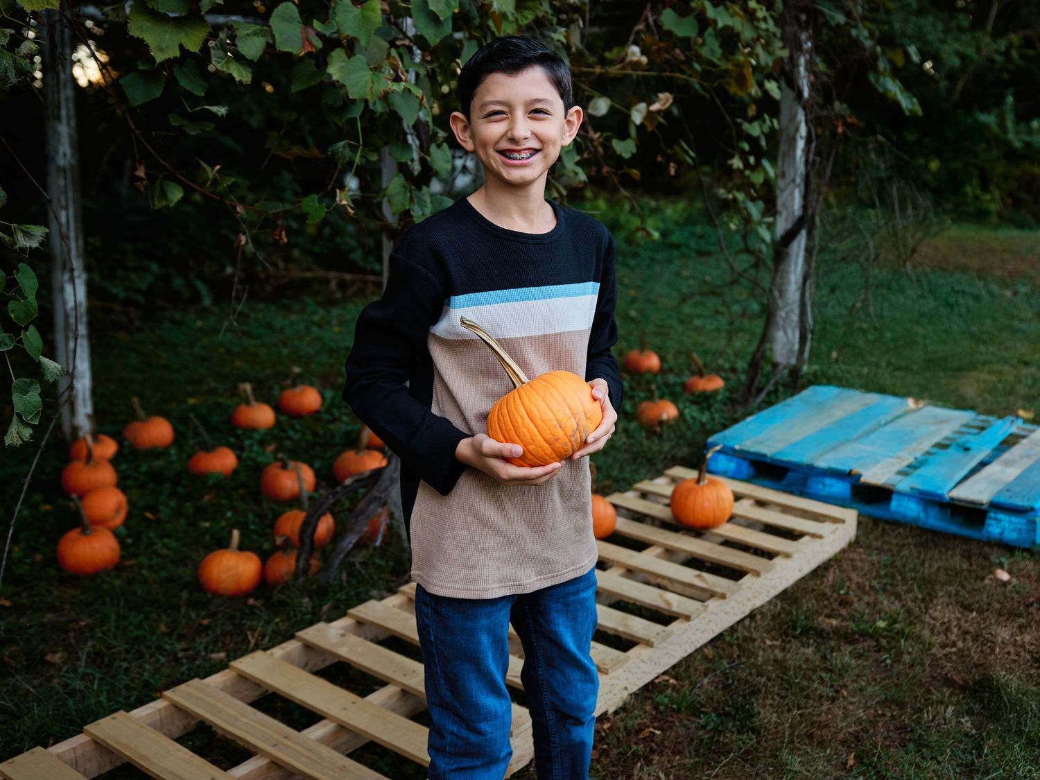 Children picking out pumpkins at a rustic outdoor pumpkin patch display with wooden pallets and fall foliage.