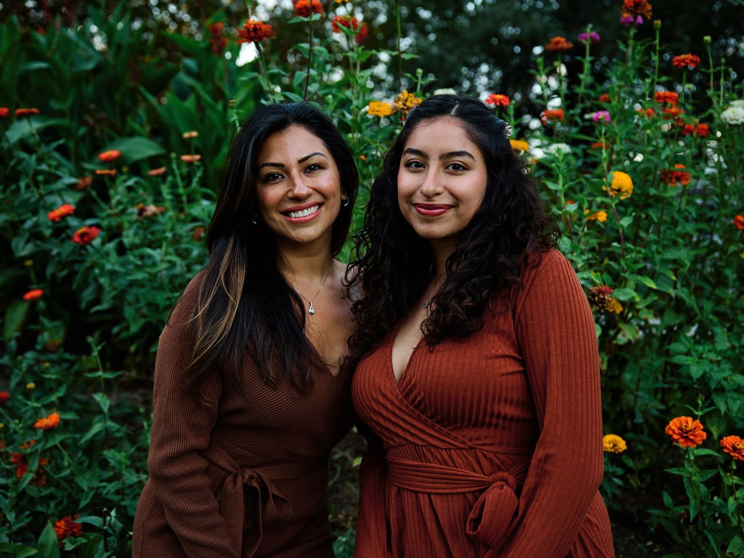 Two women in brown wrap dresses pose together in front of a colorful garden with blooming flowers.