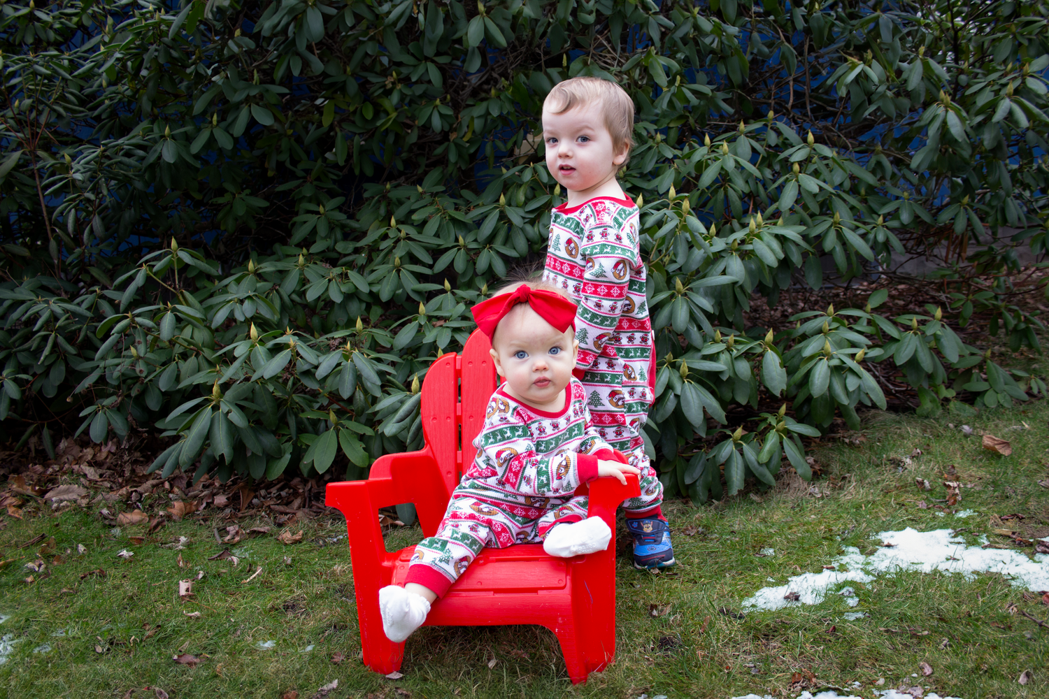 Two young children in floral outfits sitting on a red Adirondack chair in a garden.