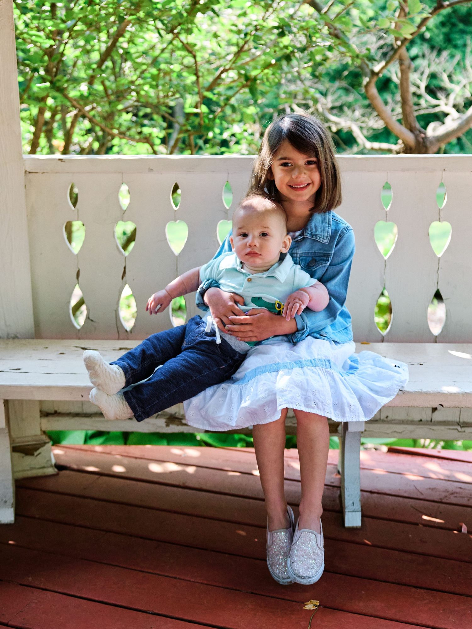 Mother and baby sit together on a wooden bench with decorative lattice railing in a lush outdoor setting.