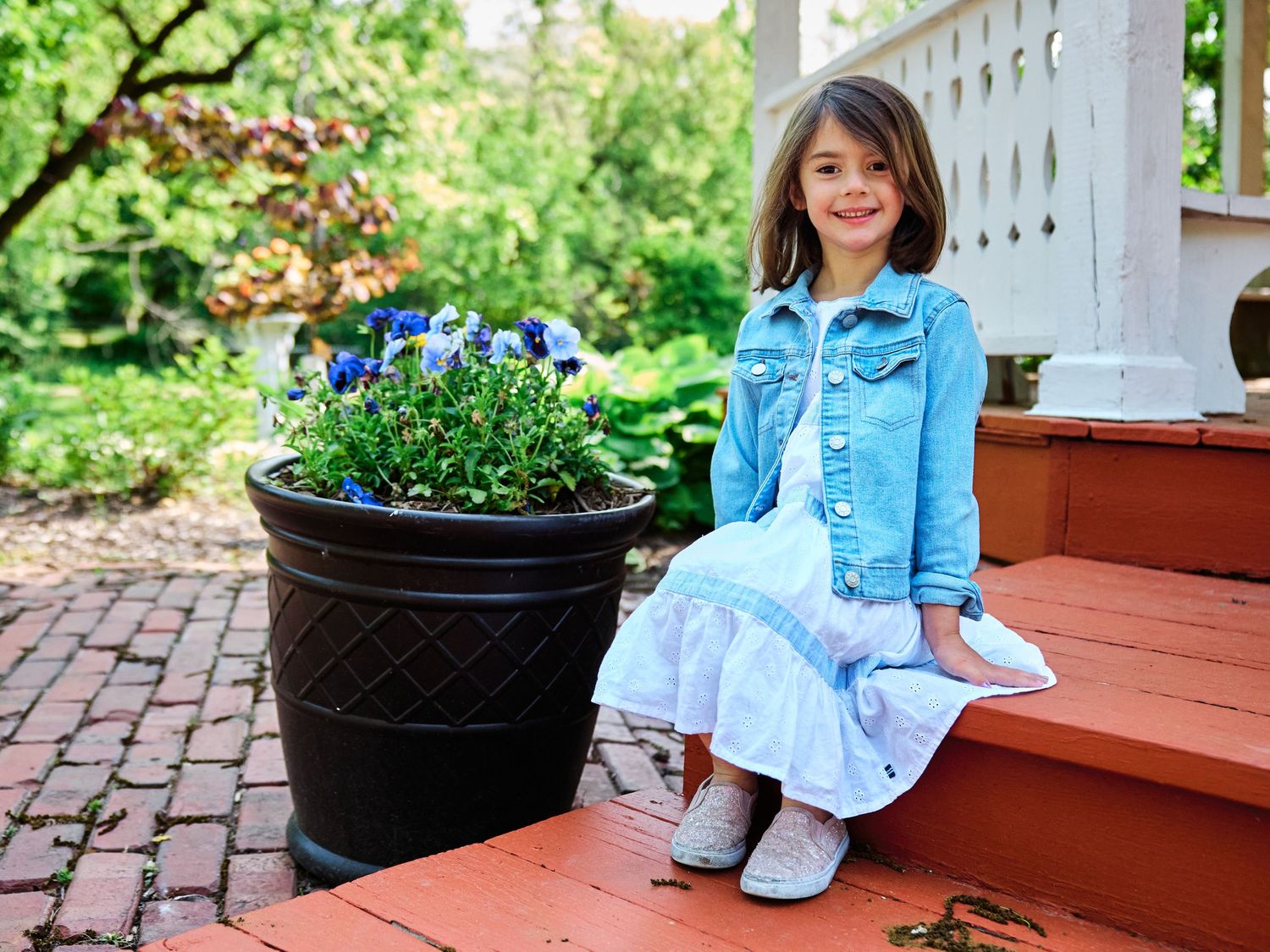 Child in denim jacket sitting on brick porch steps next to large planter with purple flowers on spring day.