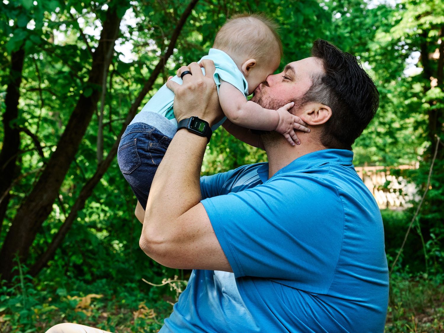 A person in blue t-shirt lifts and kisses a baby while sitting in a green forest setting.