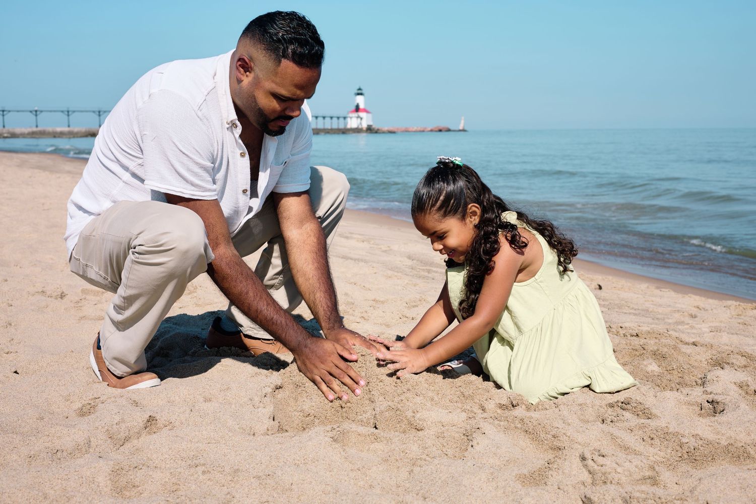 Two people building sandcastles together on a beach with a lighthouse visible in the background.