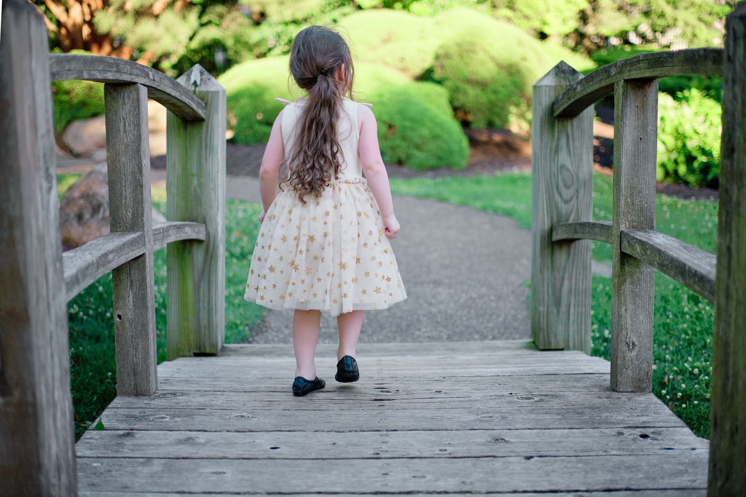 Young person in a dress walking across a wooden footbridge in a garden.