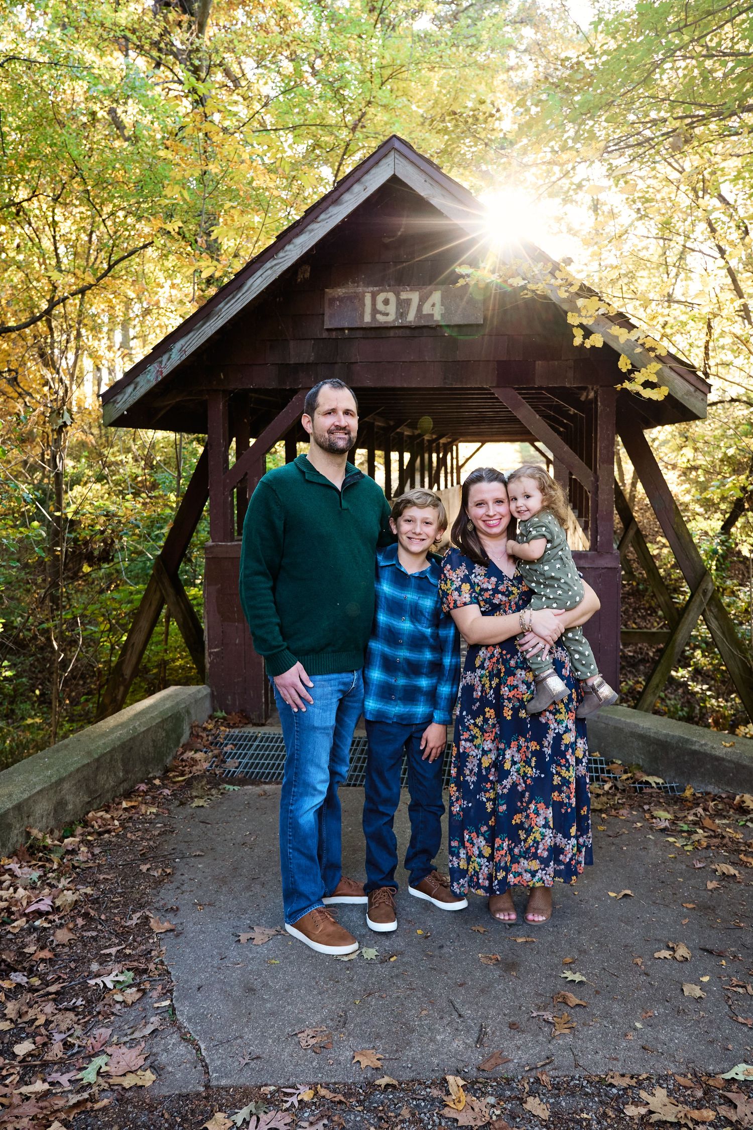 Family standing together under a covered wooden bridge dated 1974 surrounded by autumn foliage.