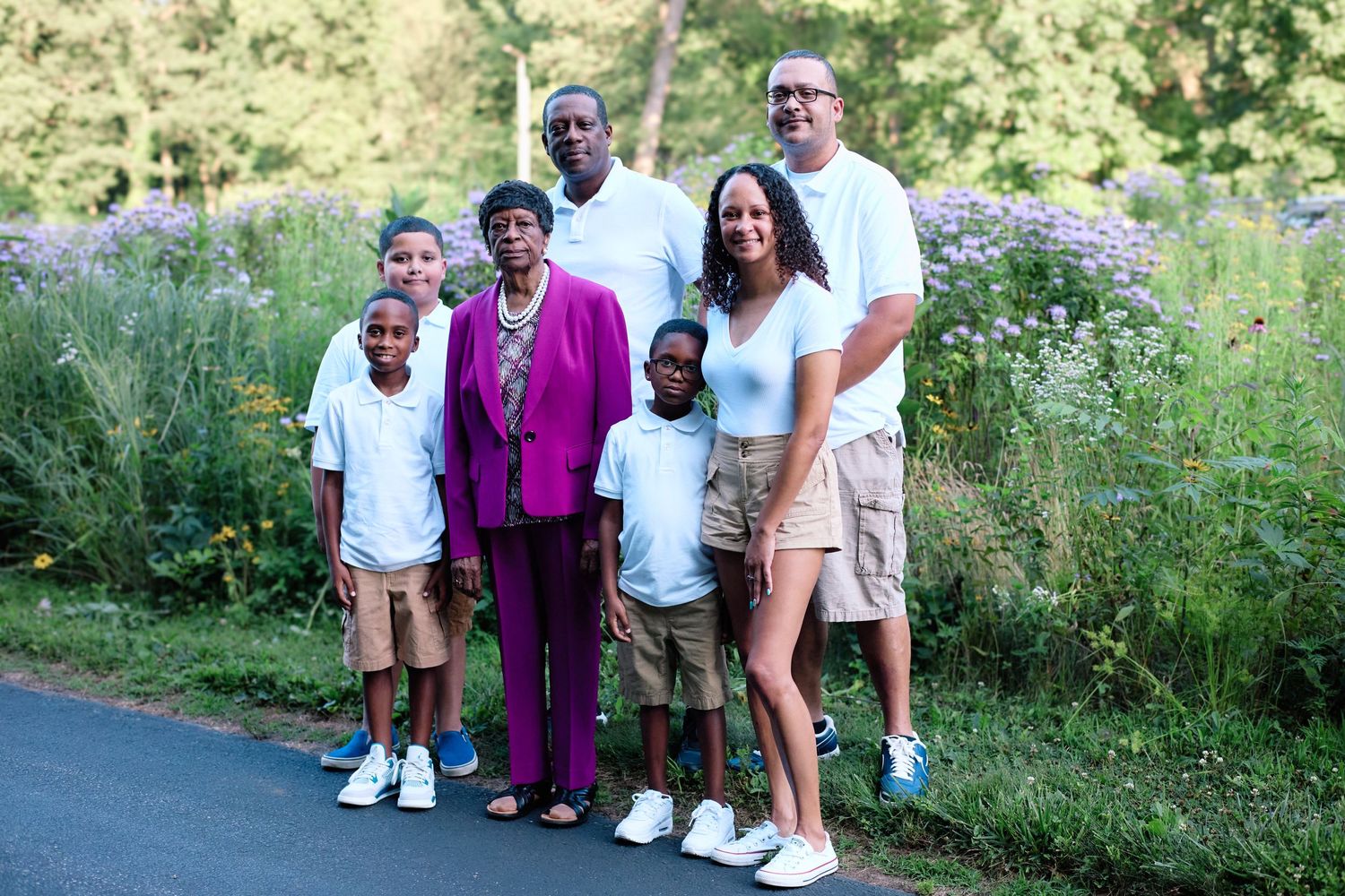 A multigenerational family stands together outdoors on a path surrounded by greenery on a sunny day.