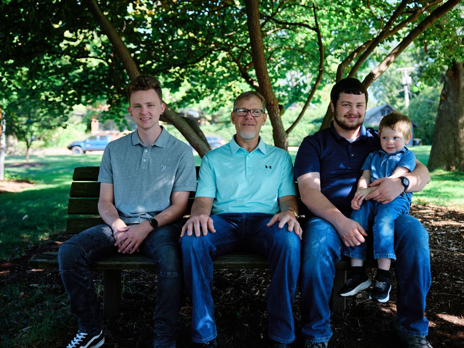 A group of people in blue jeans sit together on a wooden bench in a lush green park setting.