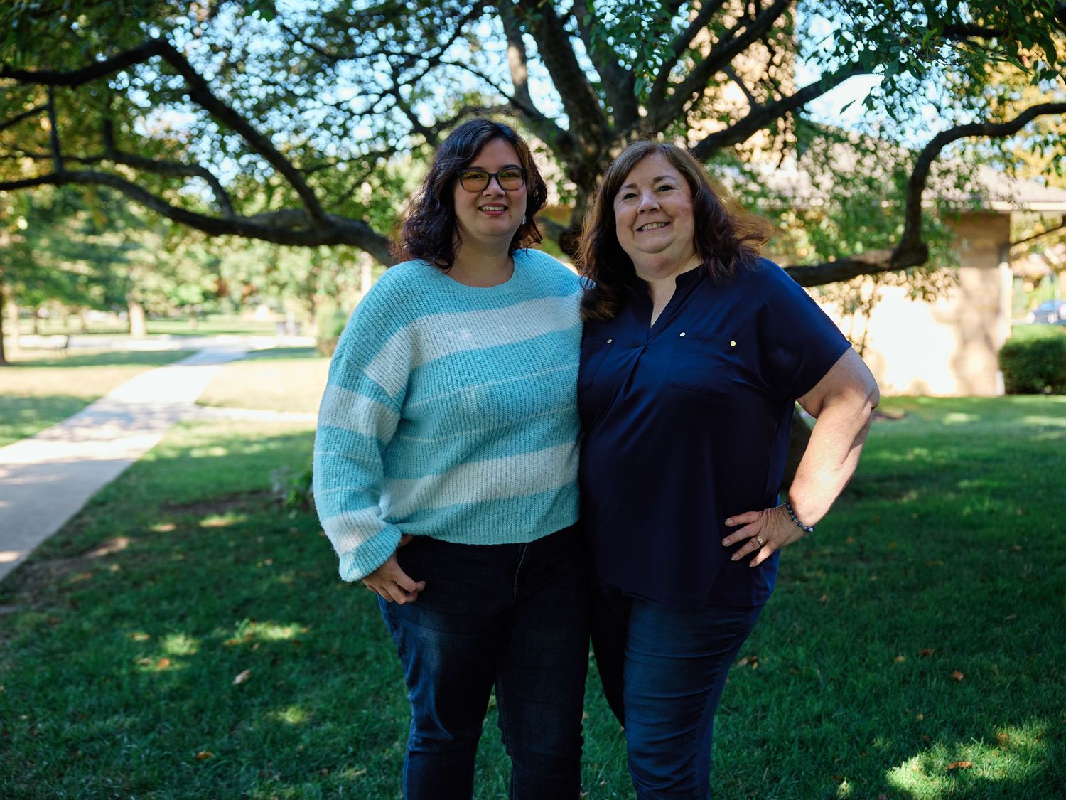 Two people stand together outdoors under green trees in a park setting during a sunny day.