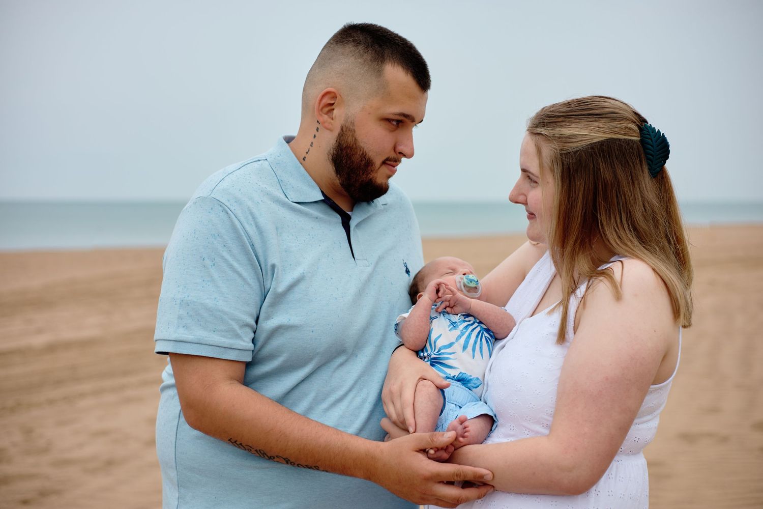 Parents holding their newborn baby at the beach during a cloudy day.