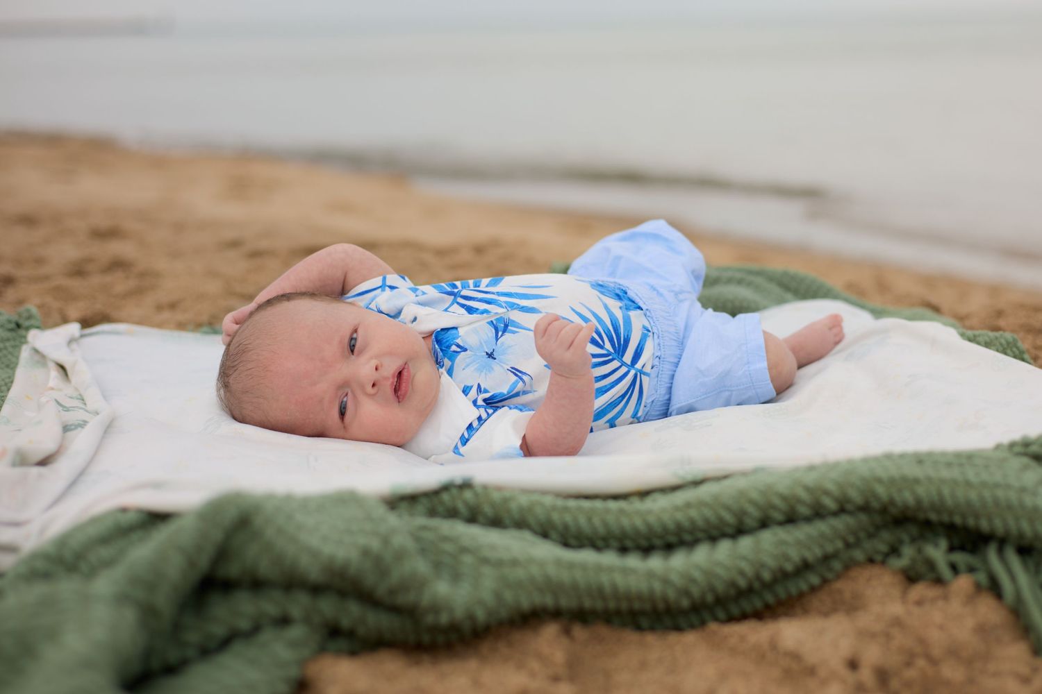 Baby in blue clothing lying on a blanket at the beach.