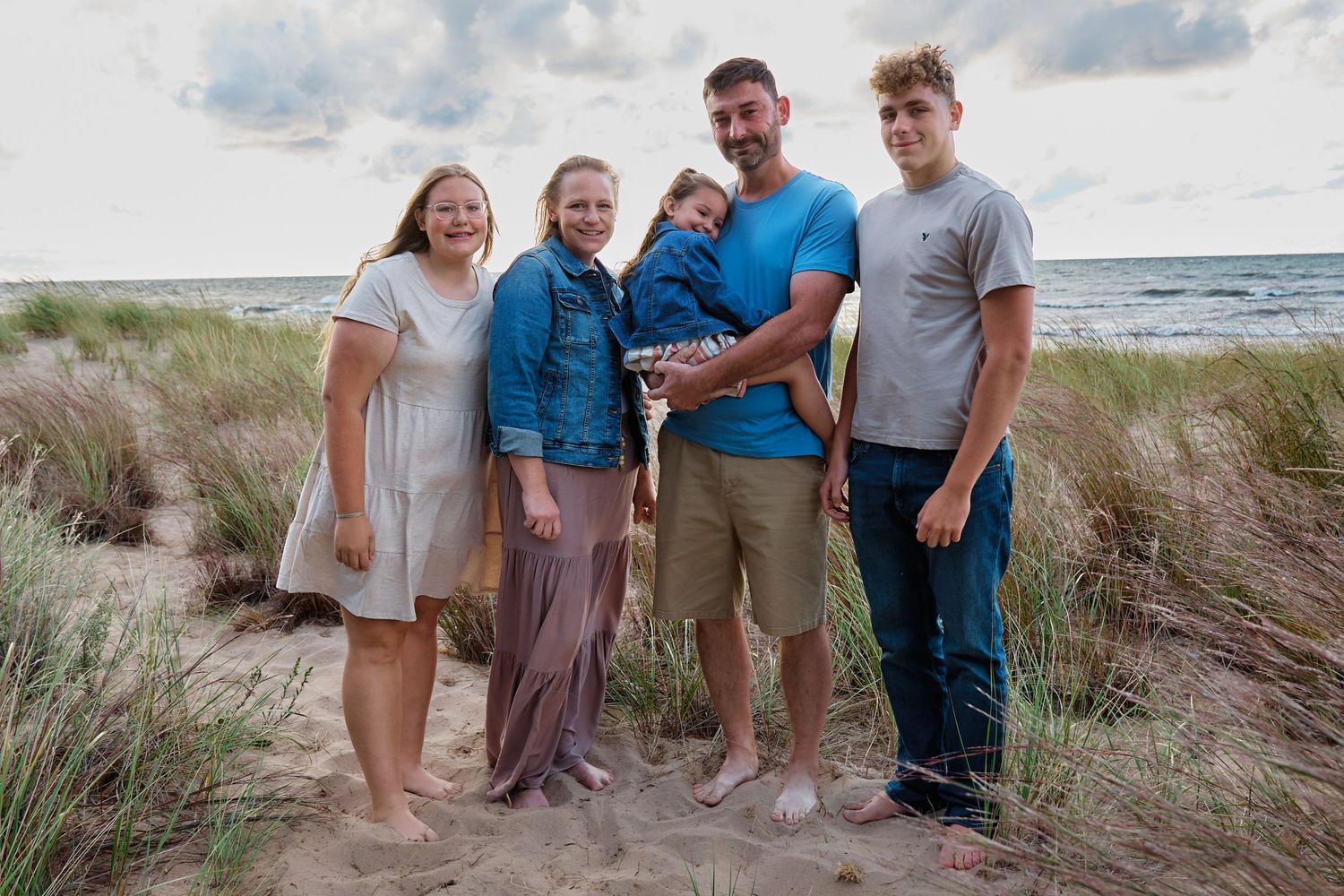 A group poses together on sand dunes with the ocean and cloudy sky in the background.