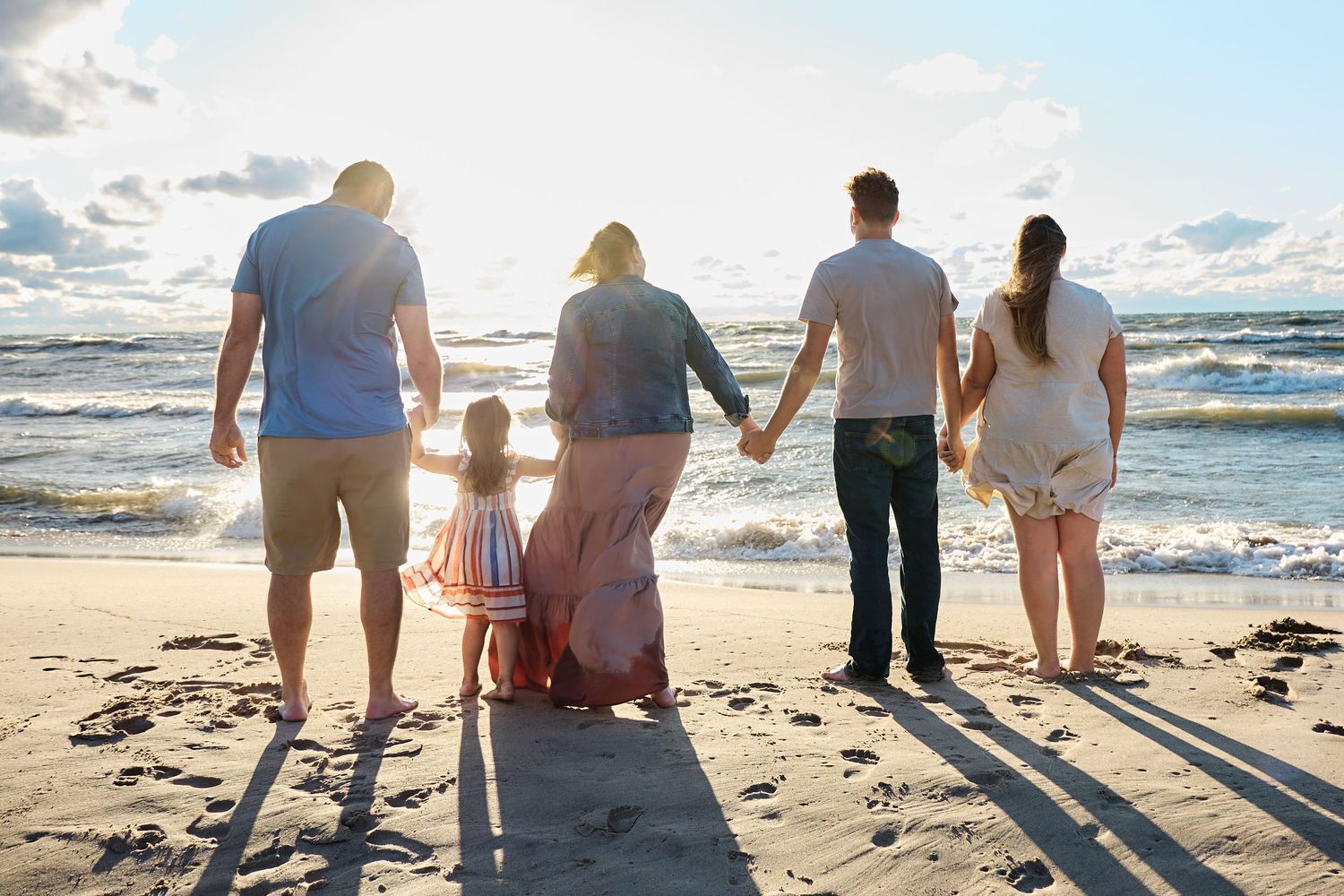 A family holds hands while walking on the beach at sunset, viewed from behind.