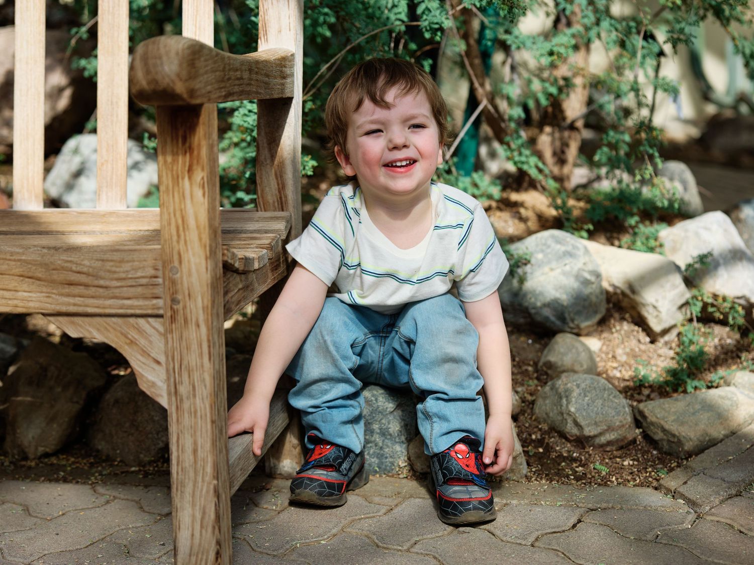 A joyful child wearing denim overalls and a stripe shirt sits on outdoor stone steps surrounded by greenery.