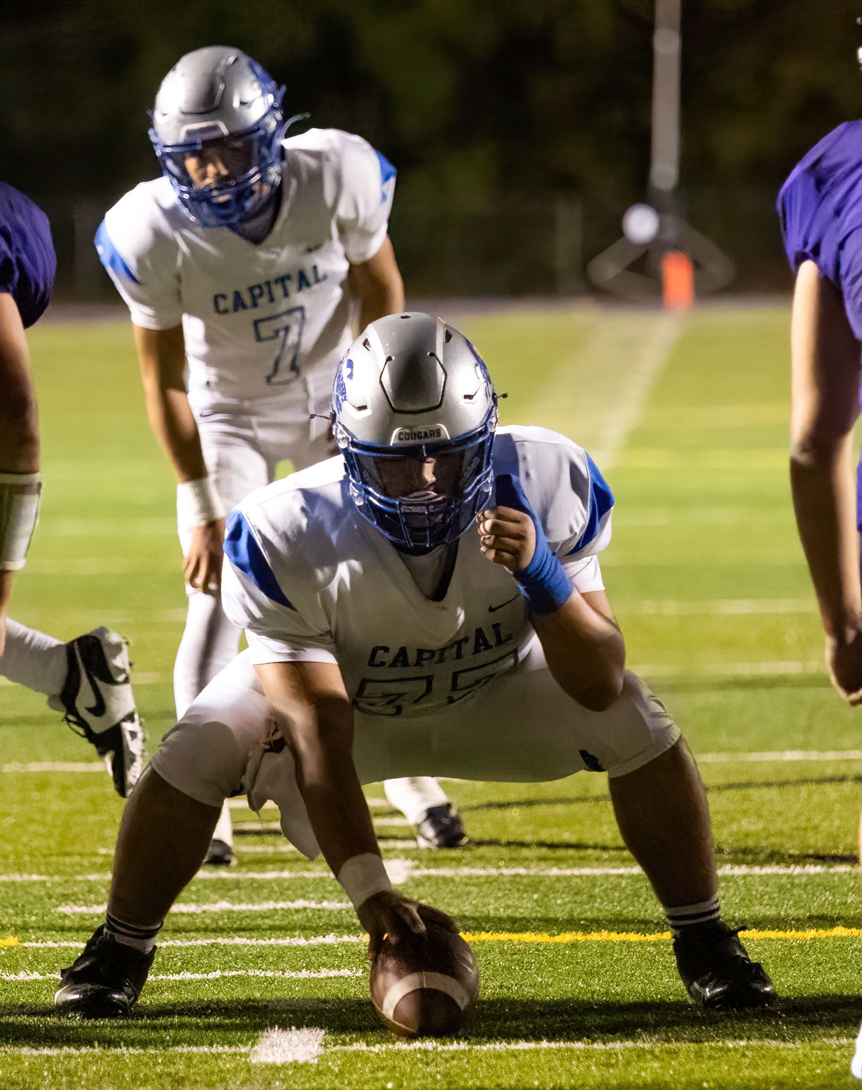 High school football player in white uniform crouches in snap position on field during nighttime game.