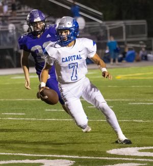 Player in white Capital uniform runs with football while being chased by defender.
