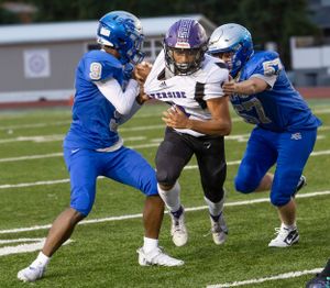 Football player in purple uniform gets tackled by two defenders in blue jerseys during game.