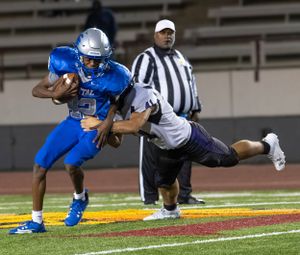 Football player in blue uniform carries ball while being tackled by defender during night game.