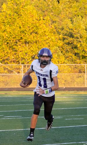 High school football player in white uniform runs with ball against autumn foliage backdrop.