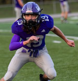 Football player in purple uniform and helmet stands ready in defensive stance.