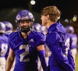 Players in purple team uniforms interact on the sidelines during a football game.