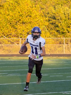 Football player in white Riverside uniform runs with ball against autumn backdrop.